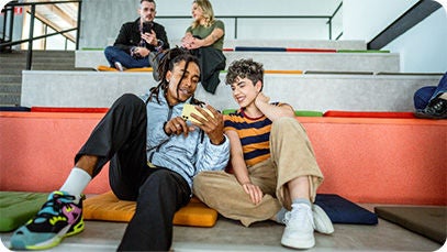 Vier Personen, zwei sitzen auf einer großen Treppe im Hintergrund, zwei sitzen in der ersten Reihe. Beide Paare nutzen ein Smartphone.