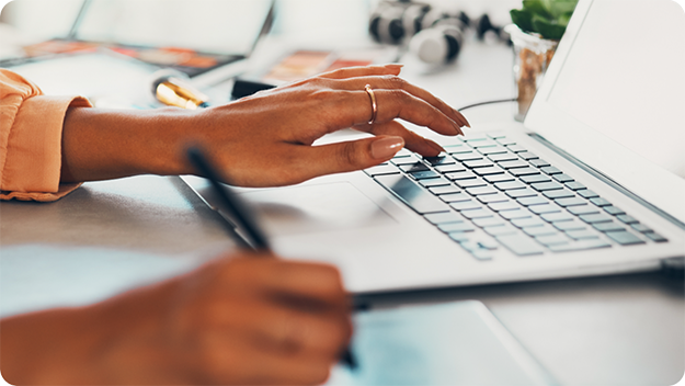 Close-up of hands working at a desk: one hand typing on a laptop keyboard, the other holding a pen over a notepad or tablet, with blurred office items like headphones and papers in the background.