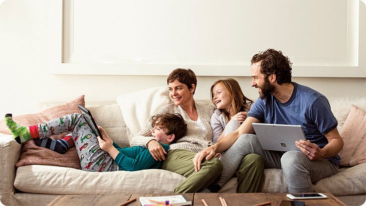 Zwei Erwachsene und zwei Kinder auf einem Sofa. Eine Person hält einen Laptop in der Hand. Ein Kind verwendet ein Tablet.