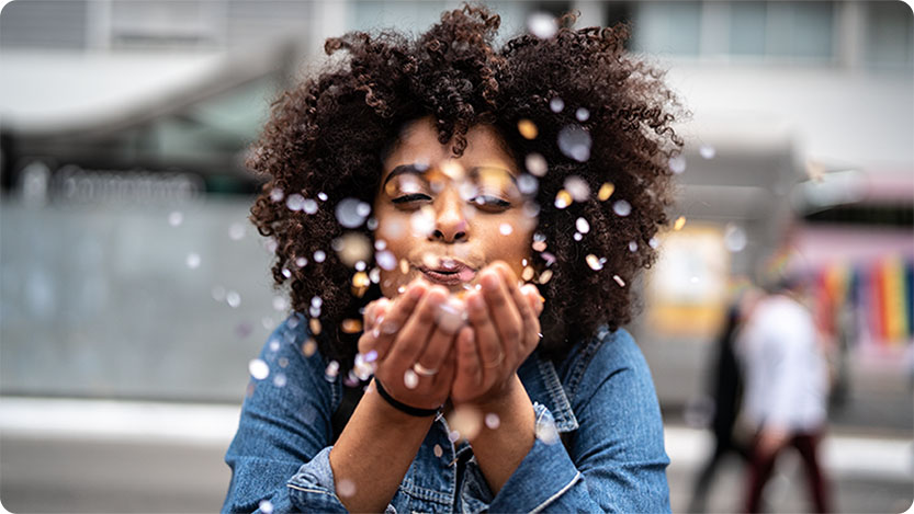 A person with curly hair wearing a denim jacket is standing outdoors, holding their hands together in front of them. Small bubbles or droplets are floating around the person's hands.