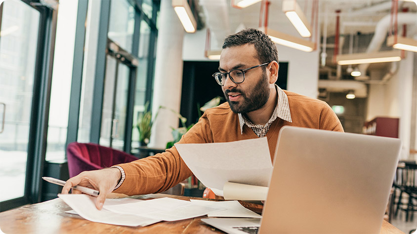 A businessperson sitting at a desk with a laptop and looking through papers
