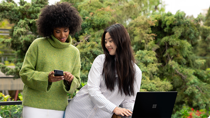 Two people standing outdoors by a railing, one using a smartphone while the other works on a laptop, with greenery in the background.